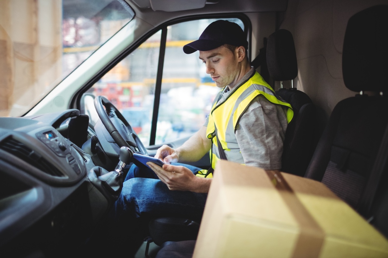 Delivery driver wearing a high-visibility vest and cap sitting in a van, using a tablet to manage shipments.