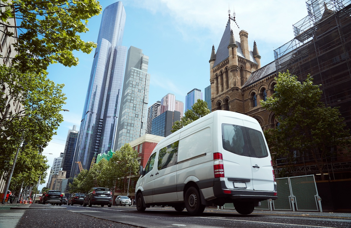 White delivery van driving through a modern city with tall skyscrapers and historic buildings.